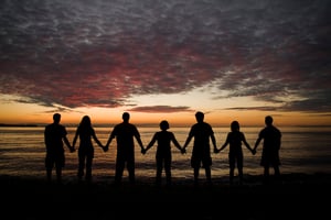 7 people holding hands on a beach, facing the sunset.