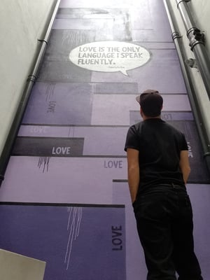 A man looking up at a part of a wall he has painted in shades of purple with the word love repeated in various places.