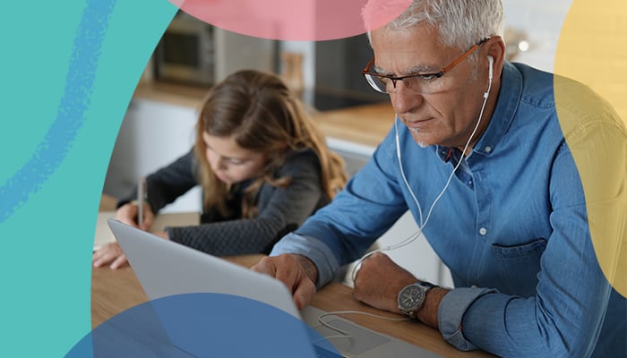 An older man wearing ear buds and working on a laptop at a table next to a little girl writing with a pencil.