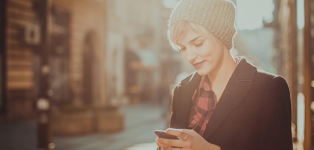 A woman standing on a sidewalk, looking at her phone. She's wearing a knit hat and a jacket.