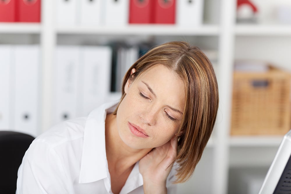 A woman in an office, leaning on her desk with her hand on the side of her neck and her eyes closed.