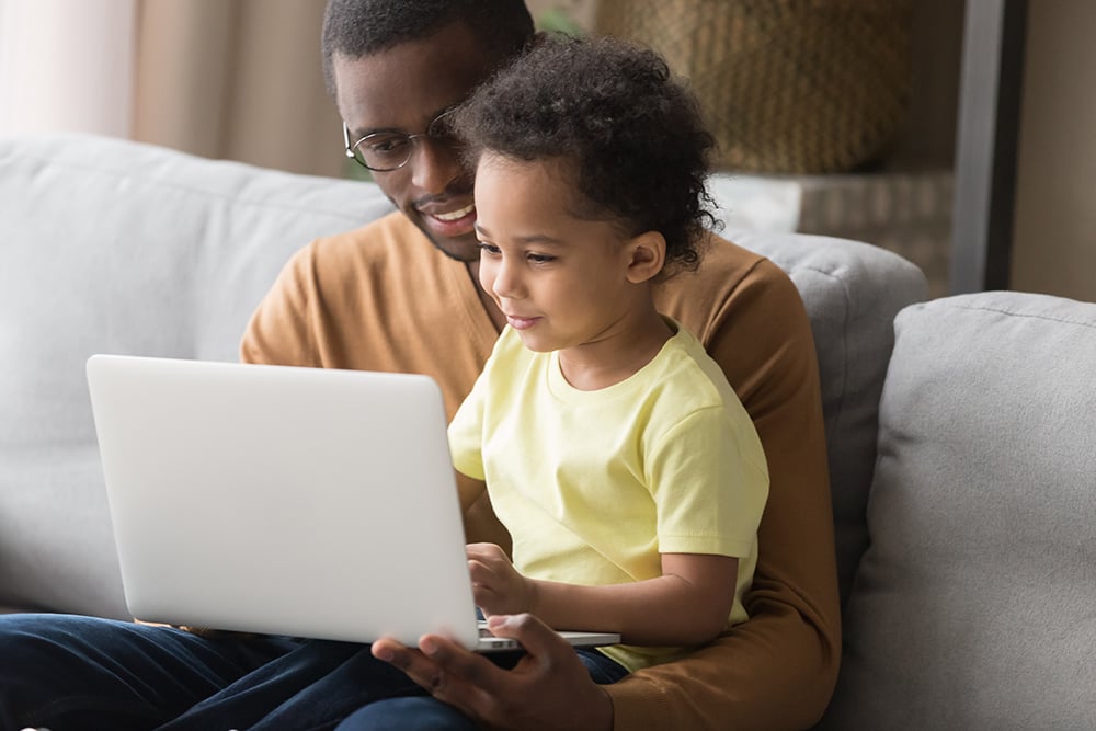 A father and young son sitting on a couch looking at a laptop together.