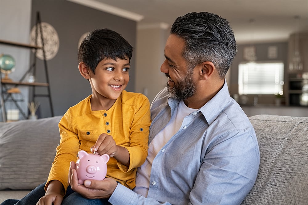 A boy sitting on her fathers lap, on a couch. The father is holding a piggy bank and the boy is putting a coin in.
