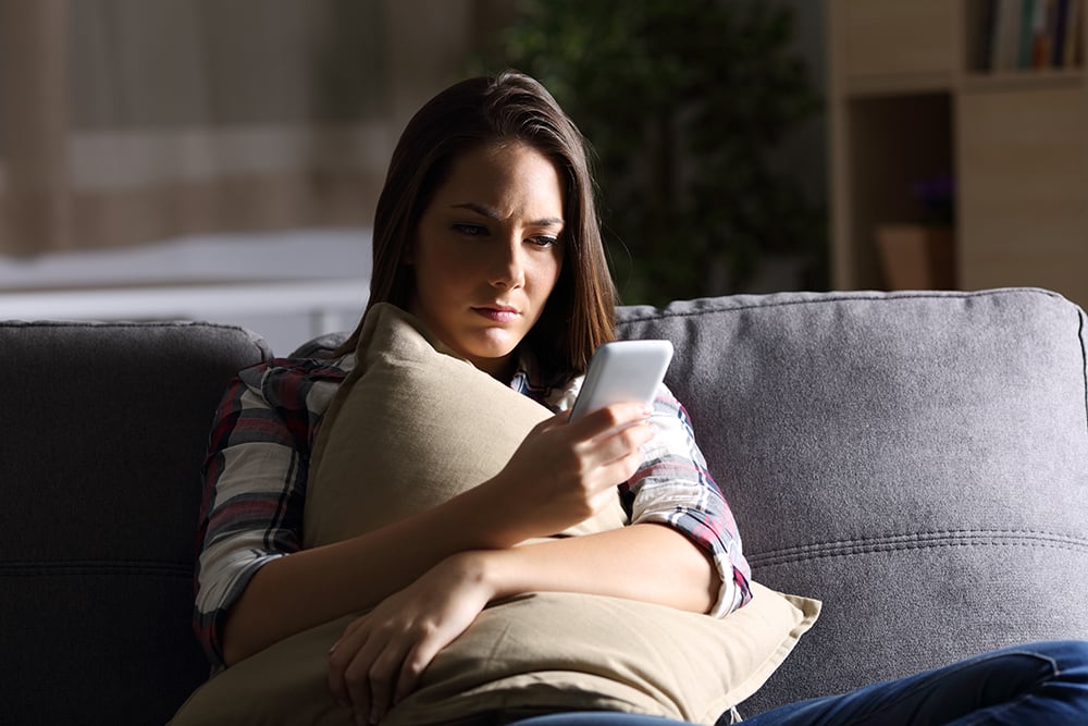 A woman sitting on a couch, holding a pillow to her body and using her phone while looking concerned.