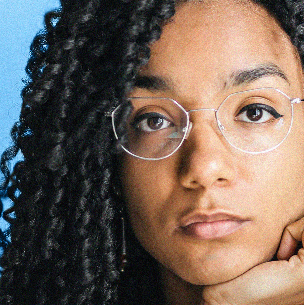 A closeup of a woman with curly black hair and glasses, looking into the camera.
